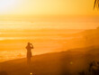 © helivideo - SILHOUETTE, LENS FLARE: Woman stands on beach and takes photo of a fiery sunset with her mobile phone. Female tourist is mesmerized by the beautiful scene at famous surf spot in Taghazout, Morocco.