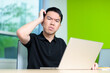 © fotolite - A young man scratches his head in confusion while looking at a laptop screen in a modern classroom. Showing problem solving, online learning frustration, or thinking through difficult tasks.