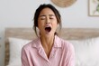 © Netsai - A young Asian woman in pink striped pajamas yawns widely with her eyes closed, looking tired in a bright bedroom.