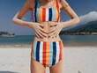 © SHOTPRIME STUDIO - Close up of a sunlit beach scene featuring a striped bikini-clad figure with hands resting on the midriff, clear water, sandy shore, and distant mountains under a bright sky.