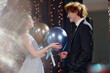 © AnnaStills - Teenage Caucasian girl smiling while handing metallic balloons to teenage Caucasian boy in formal suit, both standing close together at event decorated with string lights