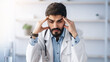 © Prostock-studio - Overwork, first symptoms of illness, migraine, feeling unwell. Tired young arabian bearded guy doctor in white coat presses hands to temples, sits at table in office interior, suffers from headache