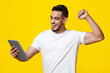 © Prostock-studio - Positive handsome millennial middle eastern man in white t-shirt trading online, enjoying success, holding digital tablet, raising hand up and smiling, yellow studio background, copy space