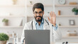 © Prostock-studio - Telemedicine, medical online, meeting with client at home during lockdown. Smiling young arabian bearded doctor in white coat, glasses looks in laptop and waves hand in office interior in clinic