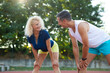 © Dexon Dee - Man and woman stretching together on running track, smiling during outdoor workout.