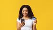© Prostock-studio - Easy money transfer. Smiling young african american lady holding debit credit card and using her smartphone. Lady making online payment standing over yellow studio background