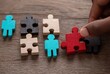 © Angra - A hand places a red puzzle piece among others of various colors and human shapes on a textured wooden surface, suggesting connection and problem solving
