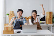 © amnaj - Two happy business owners wearing aprons are celebrating a successful online sale in their warehouse full of cardboard boxes