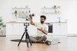 © sofiko14 - Adult man records fitness video blog at household kitchen, explaining dumbbell exercises. Sitting on floor with camera, tripod, and laptop, demonstrating healthy lifestyle ideas.