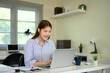 © Prathankarnpap - Young woman smiling while working on laptop at home office desk with coffee cup