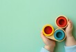 © Aliakbar - Close-up of hands holding three wooden toys in red, blue, and yellow colors on a light green background with copy space for text.