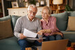 © Stockphotodirectors - An elderly couple, a man and woman, sit on a blue sofa inside a home, reviewing papers related to financial planning, insurance, and retirement near a laptop.