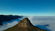 © AmazingAerialAgency - Aerial view of Lion's Head peak piercing through a sea of cloud, contrasting with the deep blue sky above, Cape Town, Western Cape, South Africa.
