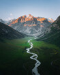 © AmazingAerialAgency - Aerial view of a winding river snaking through a vibrant green valley, framed by rugged mountains kissed with golden light, Karakol, Issyk-Kul Region, Kyrgyzstan.