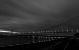 Black and white night view of the Humber Bridge illuminated with streetlights, stretching across the water under a cloudy sky with reflections on the surface below