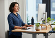 © Peeradontax - A professional woman works comfortably at a modern, adjustable standing desk in a bright office. She smiles while using her computer, showcasing an ergonomic and healthy approach to the modern workday