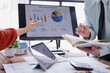 © Tj - Business colleagues discussing sales performance while reviewing charts and graphs on a computer screen, analyzing financial data in a modern office setting