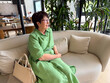 © satura_ - Middle-aged woman in green dress sitting on sofa with handbag. Lifestyle, leisure, and modern relaxation indoors.