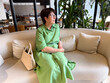© satura_ - Middle-aged woman in green dress sitting on sofa with handbag. Lifestyle, leisure, and modern relaxation indoors.