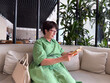 © satura_ - Middle-aged woman in green dress reading book on sofa. Knowledge, leisure, and personal time in modern lifestyle.
