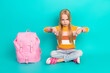 © deagreez - Young girl showing thumbs-down gesture while seated beside a pink backpack against a teal studio background