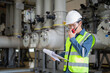 © JD Studio - Engineer in safety vest and helmet using walkie talkie during power station inspection holding documents with industrial pipes and machinery in background showing careful work
