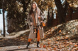 © deagreez - Chic woman in fall outfit walking through an urban park in the autumn sunshine, enjoying seasonal charm surrounded by colorful foliage