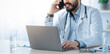© Prostock-studio - Covid-19 outbreak and medical consultation with professional. Busy young arabian bearded doctor in white coat with stethoscope sits at table with laptop, speaks on phone in office interior in clinic
