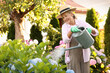 © New Africa - Smiling senior woman in hat and apron watering beautiful flowers with can in garden