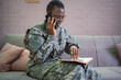 © Jelena - Smiling african american soldier talking on smartphone and reading book at home