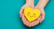 © Bhavesh - Hands holding yellow heart with smiling face on blue background, symbolizing kindness, care, happiness, love, and emotional well-being.