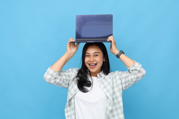 Very excited young Asian woman is lifting and showing blank laptop screen above her head, excited and cheerful, looking at camera, standing isolated on blue background.
