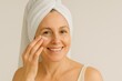 © Denis - middle aged woman looking at camera smiling with towel on head applying moisturizing cream under eyes daylight studio concept of anti aging beauty skincare