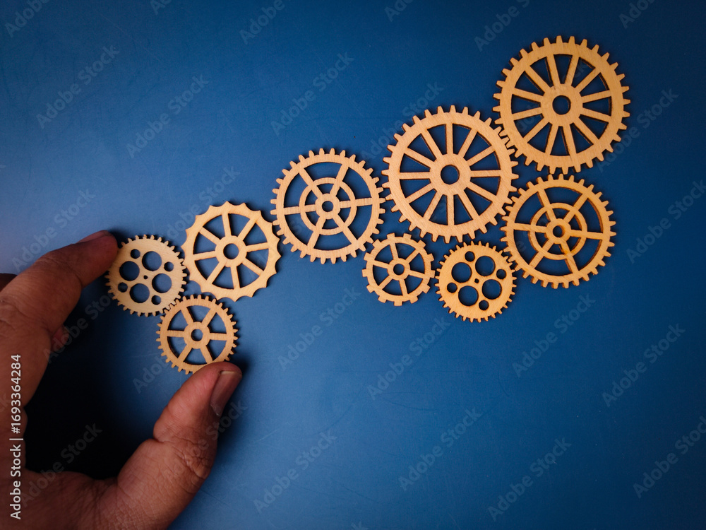 The image shows a hand placing a wooden gear, which is part of a larger ...