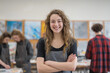 © Creative Art7 - a portrait of an high school student, smiling and standing with her arms crossed in front of art class tables filled with students working on various projects