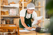 © JackF - Confident skilled young bakery manager in green apron reviewing paperwork and takes notes, organizing daily workflow in small artisan bakery