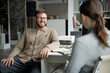 © Seventyfour - Caucasian young adult man smiling and talking with Caucasian young adult woman in modern office setting, both sitting at desk with stack of books and open notebook visible