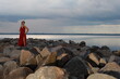 © Сергей Луговский - Pretty young brunette woman in red dress walking by rocks at the seashore