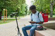 © Mediaphotos - Black young man sitting on outdoor bench using smartphone, wearing delivery backpack, electric scooter parked nearby, bicycle helmet resting on bench, summer park setting