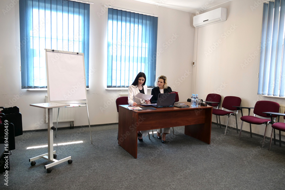 Two women at desk with laptop and papers in conference room, reviewing project documents and preparing during office teamwork session