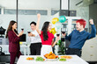 © Nuttapong punna - Energetic coworkers cheer with red wine at a lively office party table, celebrating teamwork, success, and joy with food, laughter, and festive decorations.