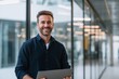 © Yuliia Malovana - Smiling businessman with beard look at the camera while standing and holding digital tablet in modern glass office. Portrait of male employee with laptop in his hands, dressed in business casual.