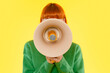 © Iryna - Young woman wearing a green sweater holds a megaphone in front of her face against a bright yellow background during a creative expression event