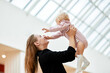 © Georgii - Young attractive mother lifts up her baby on outstretched arms in public place, shopping mall. Mom and little daughter in pink clothes play, relax and have fun while shopping. Family weekend in mall.