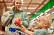 © Georgii - Happy young mother with her cute daughter in shopping trolley walking around supermarket. Beautiful mom and little blonde baby girl choosing fruit and having fun. Family shopping, happy childhood.