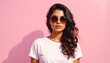 © jadid - Confident and stylish young woman with long wavy hair wearing sunglasses and a white t-shirt, posing against a solid pink background