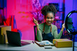 © Parichat - Young african american woman working at home selling online, recording a video blog, pointing up with a smile