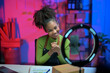 © Parichat - Young african american woman working from home selling products online using led ring light and smartphone