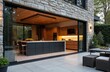 © Viktor - Modern outdoor kitchen extension features stone walls, sleek wooden cabinetry. Large island with bar stools, black counters, glass doors offers ample space. Integrated lighting, plants, patio
