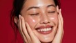 © Tina - Smiling young Asian woman with long black hair, holding her face with both hands. Bright red background enhances her joyful expression.